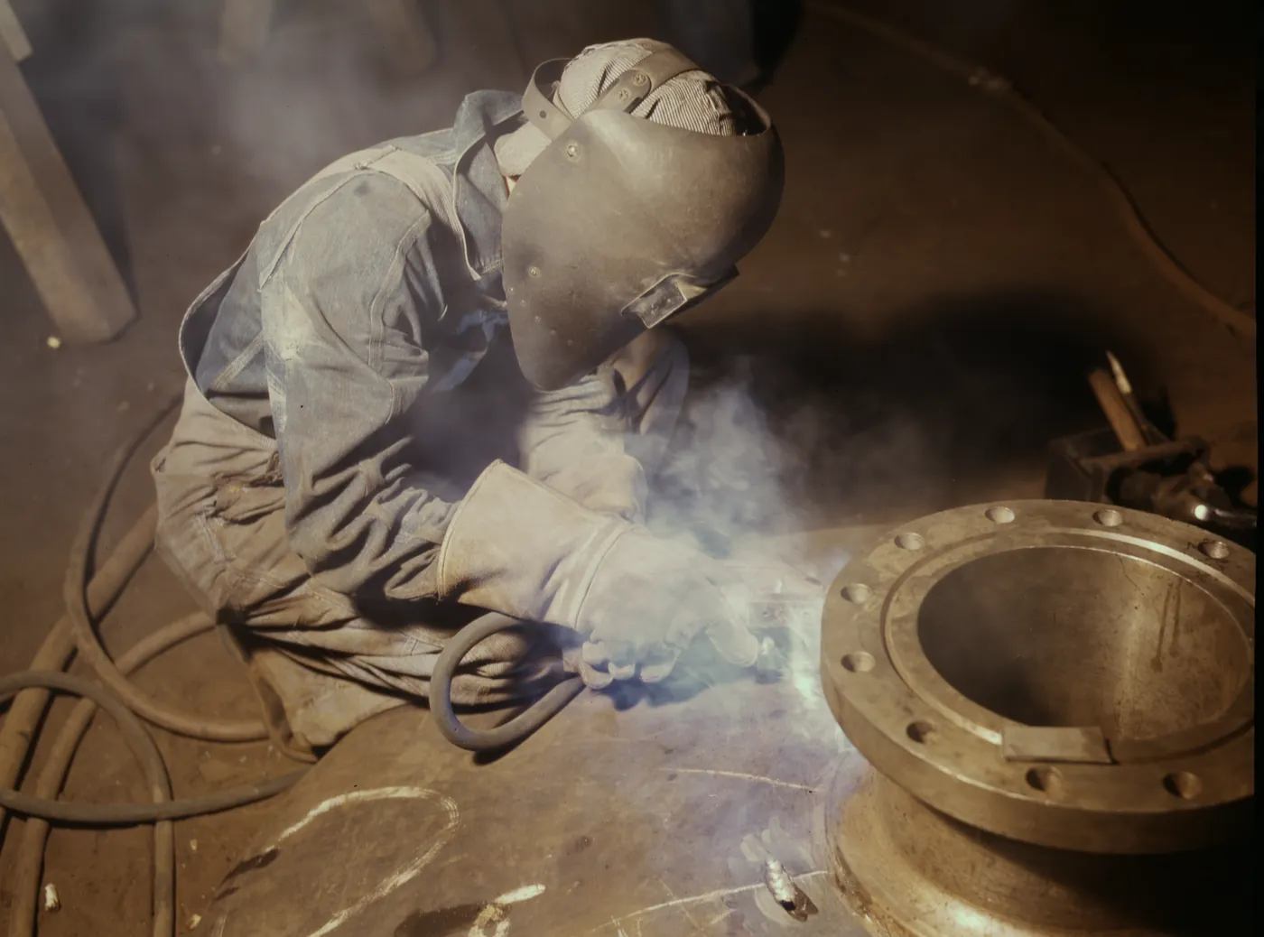 Archival photograph: a welder kneeling over a large steel flange on a shop floor, smoke rising from the joint, hose coiled beside him. Alfred T. Palmer for the Office of War Information, 1942.
