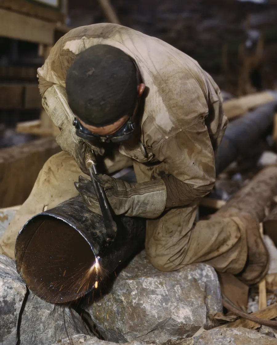 Archival photograph: a man in goggles cutting the end off a length of steel pipe with a torch, sparks running down onto the rock beneath it. Alfred T. Palmer for the Office of War Information, 1942.