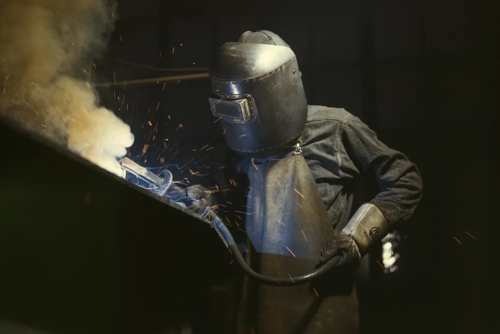 Archival photograph: a welder in a leather hood and apron running a bead on steel, sparks and smoke coming off the work. Alfred T. Palmer for the Office of War Information, 1942.