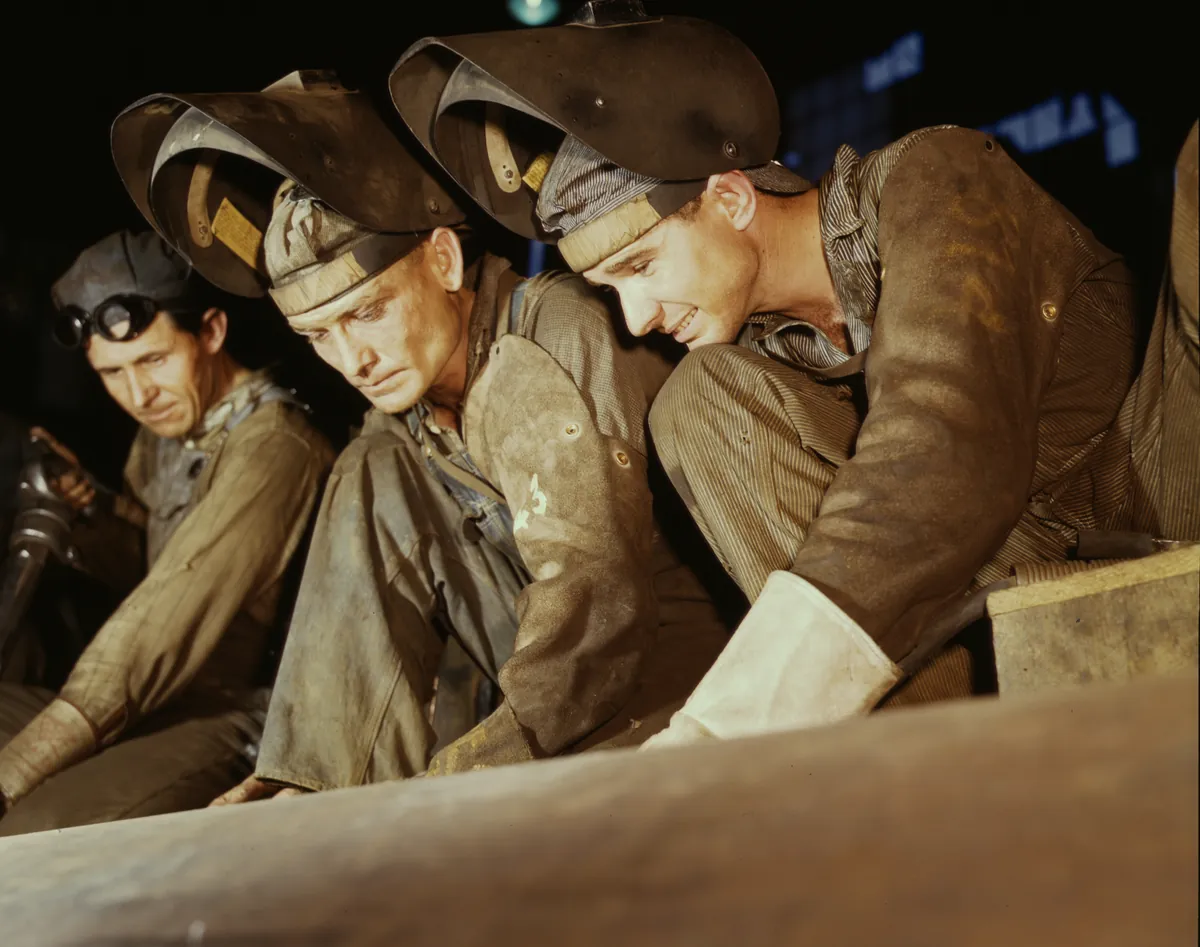 Archival photograph: three welders with their hoods flipped up, leaning over a long seam on a steel cylinder, looking at the work. Alfred T. Palmer for the Office of War Information, 1942.